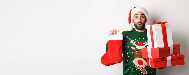 Weihnachts- und Winterferienkonzept. Aufgeregter Mann mit Weihnachtssocken und Geschenkschachteln, feiert Neujahr, bringt Geschenke unter den Baum, steht vor weißem Hintergrund. - Foto, Bild