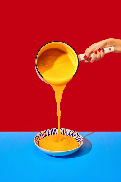 Female hand pouring delicious cheese soup into plate on blue tablecloth over red background. French dinner. Food pop art photography. Complementary colors. Copy space for ad, text - Photo, image