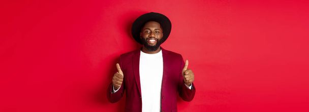 Christmas shopping and people concept. Handsome Black man smiling satisfied, showing thumbs-up, like and agree, approve something, standing against red background. - Photo, image