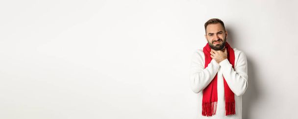 Зима, ковид-19 и концепция здоровья. Больной человек жалуется на боль в горле, трогательную шею и гримасы от боли, стоя в свитере и красном шарфе, белый фон. - Фото, изображение