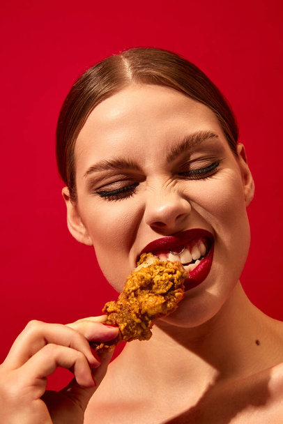 Young woman with red lipstick eating fried chicken, nuggets over vivid red background. Fast food lover. Food pop art photography. Complementary colors. Junk food. Copy space for ad, text - Photo, image
