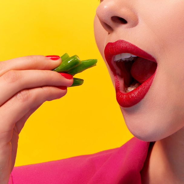 Zugeschnittene Nahaufnahme einer jungen Frau mit rotem Lippenstift, die gekochte grüne Bohnen vor gelbem Hintergrund isst. Ernährung und Gesundheit. Food Pop Art Fotografie. Komplementärfarben. Kopierraum für Werbung - Foto, Bild