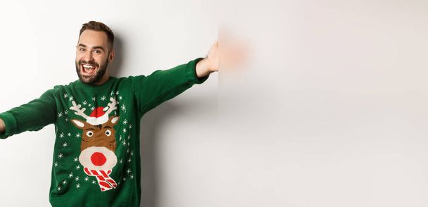 Neujahrsfeier und Winterferienkonzept. Freundlicher Mann spreizt Hände zur Umarmung, willkommen zur Weihnachtsfeier, trägt lustigen Pullover, weißer Hintergrund. - Foto, Bild