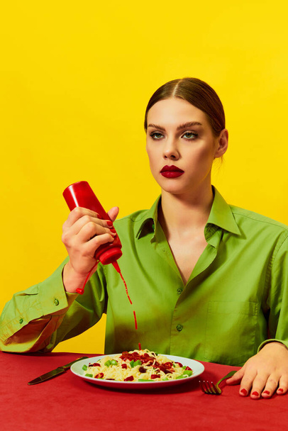 Young girl with emotionless face pouring ketchup on plate with spaghetti on red tablecloth over yellow background. Spicy. Food pop art photography. Complementary colors. Copy space for ad, text - Фото, изображение