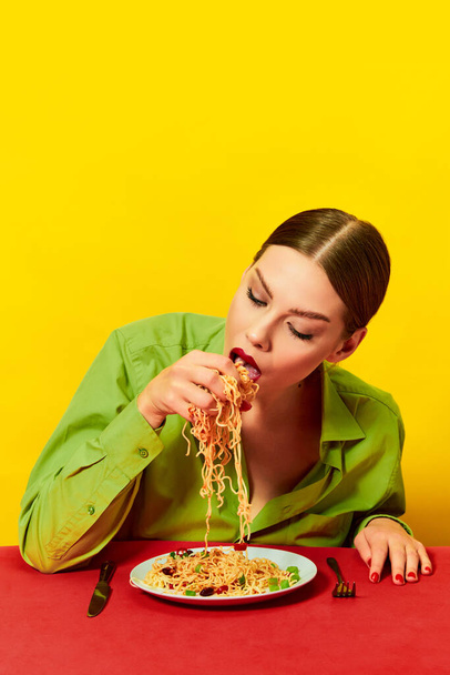 Emotional young girl eating spaghetti, noodles with hands on red tablecloth over yellow background. Delicious Italian food. Food pop art photography. Complementary colors. Copy space for ad, text - Foto, Imagem