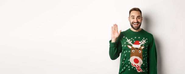 Neujahr, Feiertage und Feiern. Freundlicher junger Mann winkt mit der Hand und grüßt die Leute auf der Weihnachtsfeier, grüßt, steht über weißem Hintergrund. - Foto, Bild