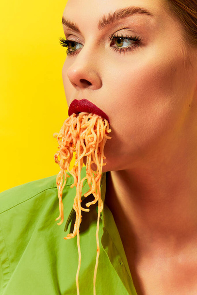 Close-up image of young girl eating spaghetti, noodles sticking out of the mouth over yellow background. Vintage style. Food pop art photography. Complementary colors. Copy space for ad, text - 写真・画像