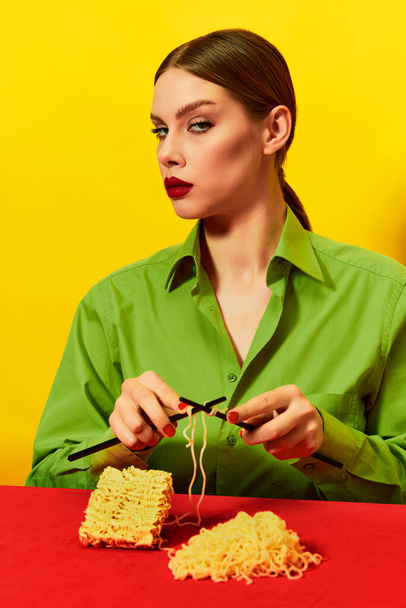 Woman with emotionless face knitting instant noodles on blue table on vivid red tablecloth over yellow background. Food pop art photography. Complementary colors. Copy space for ad, text - Photo, Image