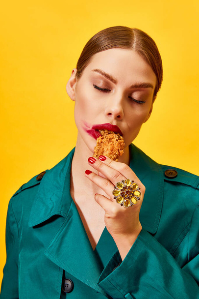 Young woman in green coat with red lipstick smudge, eating fried chicken, nuggets over yellow background. Junk food lover. Food pop art photography. Complementary colors. Copy space for ad, text - Photo, Image