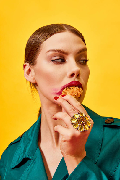 Young woman in green coat with red lipstick smudge, eating fried chicken, nuggets over yellow background. Fast food. Food pop art photography. Complementary colors. Copy space for ad, text - Photo, Image