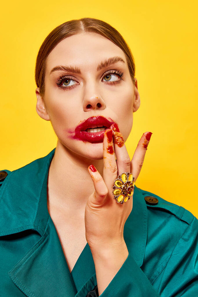 Young beautiful woman in green coat with smudged red lipstick and dirty fingers after eating nuggets over yellow background. Food pop art photography. Complementary colors. Copy space for ad, text - Photo, image