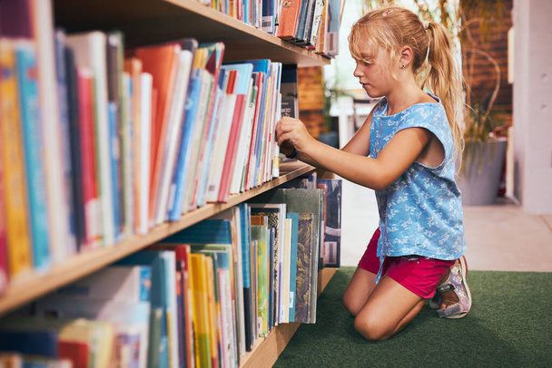 Schoolgirl looking for book for reading in school library. Student selecting literature for reading. Books on shelves in bookstore. Learning from books. Back to school. Elementary education. Doing homework - Φωτογραφία, εικόνα