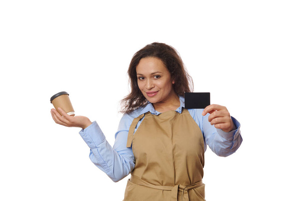 Attractive waitress bartender, barkeeper in beige chef apron, smiles looking at camera, holds a cardboard cup of hot drink to go and plastic bank card, isolated over white background. Food and drink - Φωτογραφία, εικόνα