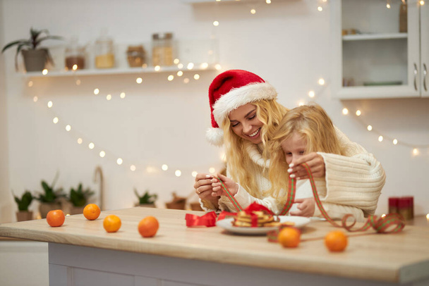 Side view of happy family wearing knitted sweaters, posing indoors, at decorated, cozy studio. Blonde woman wearing christmas hat sitting with small girl. Concept of winter holiday. - Valokuva, kuva