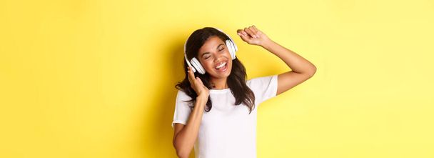 Χαρούμενη έφηβη Αφρο-Αμερικανίδα που ακούει μουσική με ακουστικά, χορεύει αισιόδοξη και τραγουδάει, στέκεται πάνω από κίτρινο φόντο. - Φωτογραφία, εικόνα