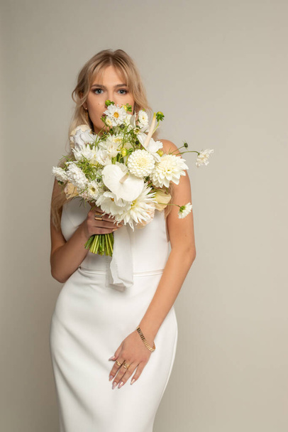 Vertical confident, charming, romantic blond female bride in white elegant dress covered by bouquet of field flowers, looking at camera in studio. Engagement and marriage concept. Copy space - Foto, afbeelding