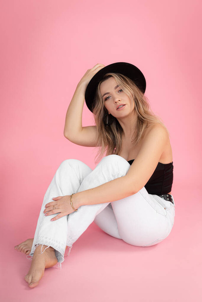 Vertical portrait of young barefoot charming, coquettish blond long haired woman in black hat sitting on floor with crossed legs and look at camera on pink background. Black and white. Copy space - Photo, image
