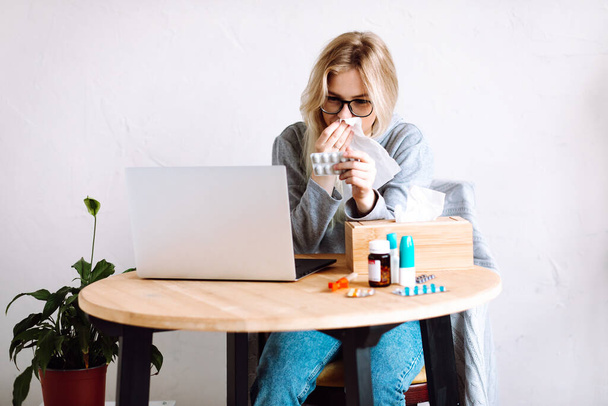 Portrét mladé ženy sedící u notebooku, láhve s modrými uzávěry, dřevěná krabice s papírovými ubrousky, ukazující pilulky na obrazovku, otírající nos, kýchání s videohovory online. - Fotografie, Obrázek