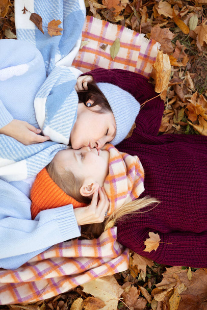 Vertical pretty, tender, loving young mother kissing gently her little blond daughter in warm clothes, lying together, stroking on foliage in nature. Child care, offspring growth. Top view, high angle - Фото, изображение