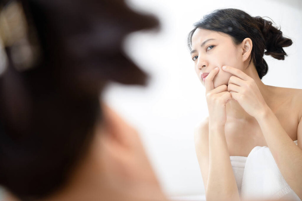 Problem skin. Concerned young asian women popping pimple on cheek while standing near mirror in bathroom. Unhappy girl inspecting face, suffering acne, focus on reflection - Photo, Image