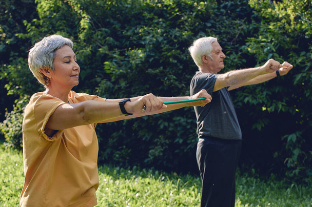 Senior 60 spouses wear sportswear doing hands exercises, training arms outdoor in summer park in the morning using resistance rubber bands. Healthy lifestyle, active retired life use modern appliance - Fotografie, Obrázek