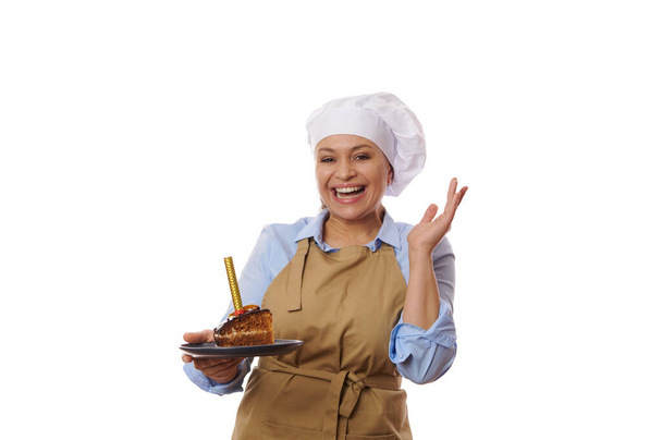 Ευτυχισμένη γυναίκα, νοικοκυρά σε chefs καπέλο και ποδιά, χαμογελώντας ένα χαρούμενο χαμόγελο toothy στην κάμερα, ποζάροντας με ένα κομμάτι σπιτικό νόστιμο κέικ σοκολάτας με αφρώδη κεριά, απομονωμένη σε λευκό φόντο - Φωτογραφία, εικόνα
