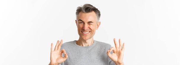 Close-up of handsome and confident middle-aged man, showing okay signs and winking, praising good choice, standing over white background. - Zdjęcie, obraz
