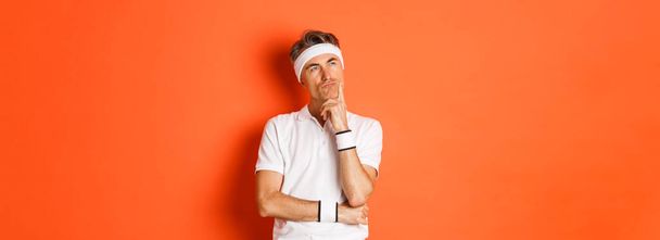 Concept of workout, sports and lifestyle. Portrait of thoughtful middle-aged man in fitness clothes, thinking about something, making decision over orange background. - Photo, Image