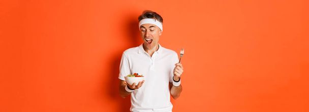 Fogalom a sport, a fitness és az életmód. Portré vidám középkorú srác edzőruhában, kezében villa és saláta, eszik egészséges ételek, álló felett narancs háttér. - Fotó, kép