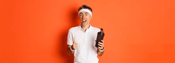 Portret van gezonde en actieve fitnessman van middelbare leeftijd, in fitnessuniform, met duimen omhoog, drinkwater, over oranje achtergrond. - Foto, afbeelding