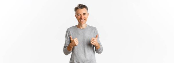 Portrait of handsome middle-aged guy with grey hairstyle, showing thumbs-up and smiling, approve something good, like idea, standing over white background. - Photo, Image