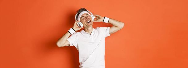 Portrait of carefree middle-aged male athlete, listening music in headphones during fitness training, standing over orange background. - Photo, Image