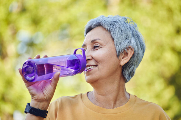 Close up image attractive mature woman holds plastic reusable bottle make sip drinking still mineral water during morning work out or stroll in summer park, caring about health, relieving thirst - Фото, зображення