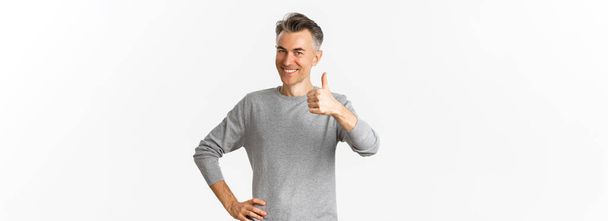 Portrait of proud and happy middle-aged man, smiling pleased and showing thumbs-up, praise something good, standing over white background. - Photo, Image