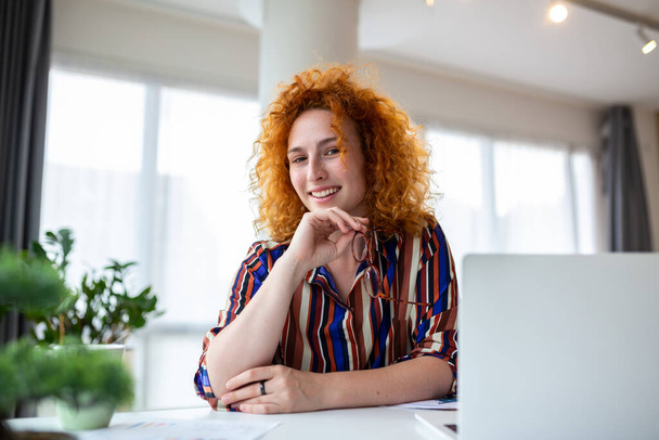Съемка улыбающейся деловой женщины, работающей с ноутбуком, глядя на камеру в современном офисе стартапа. - Фото, изображение