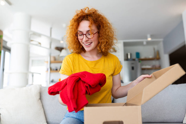 Hermosa joven alegre pelo rojo mujer unboxing un paquete en la sala de estar de un nuevo suéter. - Foto, Imagen