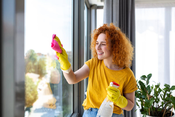 housework and housekeeping concept - happy woman in gloves cleaning window with rag and cleanser spray at home - Photo, Image