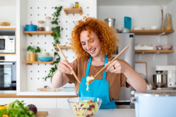 Красивая милая молодая улыбающаяся женщина на кухне готовит веганский салат в повседневной одежде
. - Фото, изображение