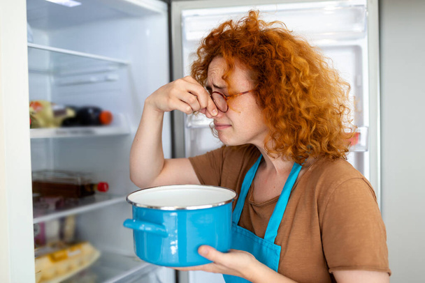 Плохая еда в холодильнике, молодая женщина держит нос из-за неприятного запаха от еды в холодильнике дома - Фото, изображение