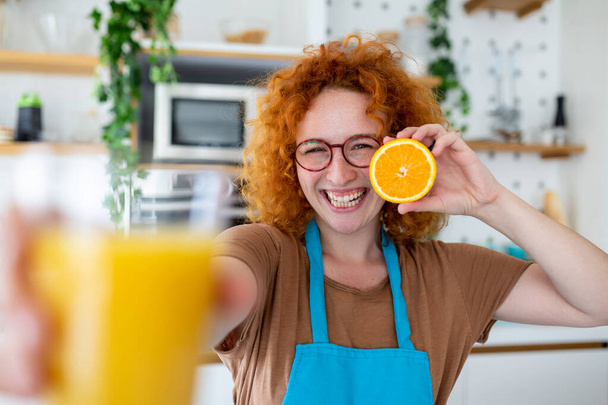 Photo of cute caucasian woman smiling and holding two orange parts while cooking vegetable salad in kitchen interior at home - Photo, Image