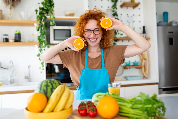 Photo of cute caucasian woman smiling and holding two orange parts while cooking vegetable salad in kitchen interior at home - Fotó, kép