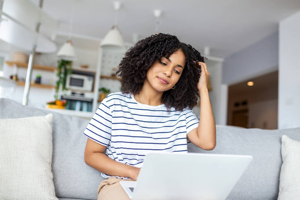 Smiling young woman freelancer working from home, sitting on couch, using laptop, copy space. Relaxed african american lady enjoying her weekend, surfing on internet on laptop - Foto, Imagen