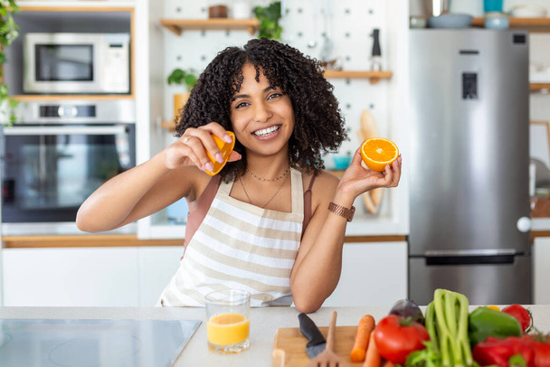 Piękna młoda kobieta pijąca świeży sok pomarańczowy w kuchni. Zdrowa dieta. Szczęśliwa młoda kobieta ze szklanką soku i pomarańczy przy stole w kuchni. - Zdjęcie, obraz