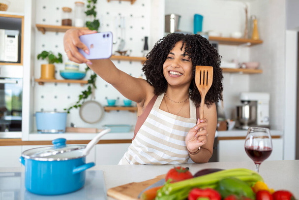 Porträt einer lächelnden jungen Afroamerikanerin, die ein Selfie mit ihrem Smartphone macht, während sie zu Hause in der Küche kocht - Foto, Bild