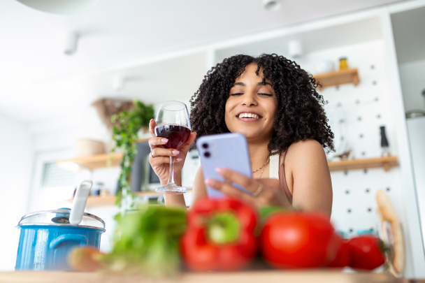 Vrouw met mobiele telefoon en een glas wijn in de keuken thuis - Foto, afbeelding