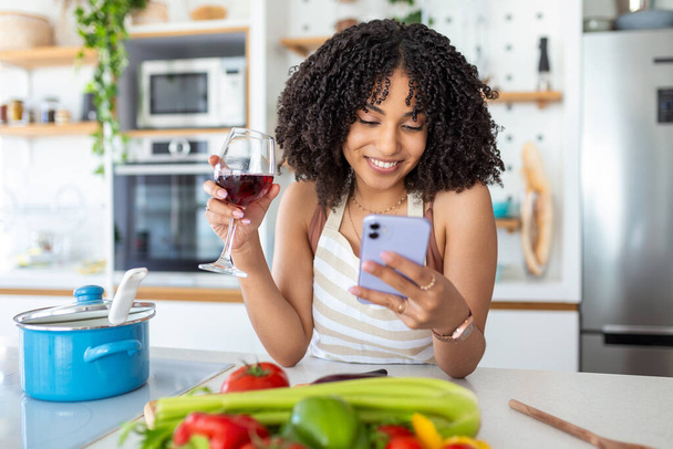 携帯電話を持っている女性と家のキッチンでワインのガラス - 写真・画像