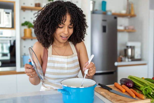 Foto de una bonita mujer afroamericana sosteniendo cuchara de cuchara mientras cocina sopa con verduras frescas en la cocina en casa - Foto, Imagen