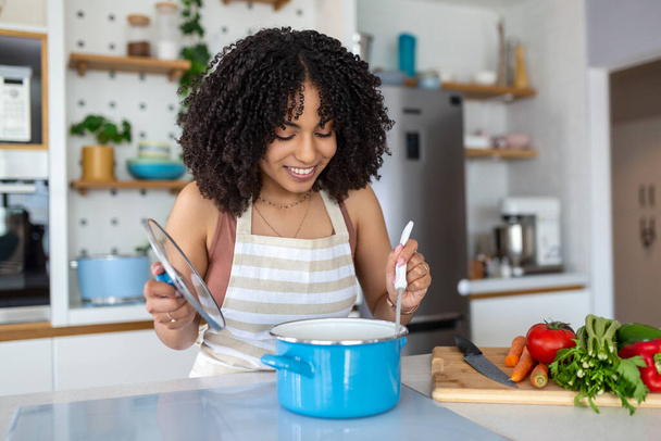 Gezond eten thuis. Gelukkige vrouw bereidt de juiste maaltijd in de keuken. Mooie glimlachende jonge vrouw in schort staan in de keuken en roeren soep in pot. - Foto, afbeelding