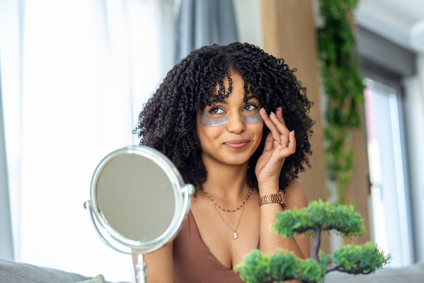 Nowoczesna kosmetologia i plastry przeciw zmęczeniu pod oczami. Wesoła młoda Afroamerykanka nakładająca je pod oczy. - Zdjęcie, obraz