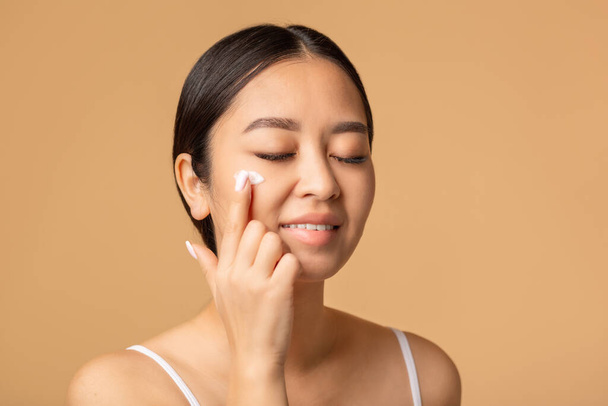 Beautiful korean woman applying a anti age or moisturizer face cream on her face, smiling sweetly standing on a beige background - Foto, imagen